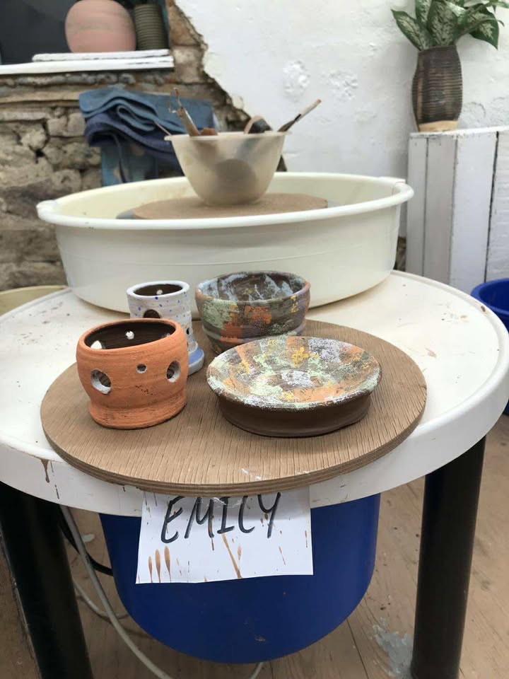Four finished pots on a circular wooden board, in front of a white pottery wheel. There is a plastic bowl on the wheel that contains a selection of pottery tools. The finished pots are as follows: a tea light pot with holes cut around the side, a smaller light blue and white pot behind (slightly hidden) and two pots with a stippled sponge surface of green, orange and yellow.