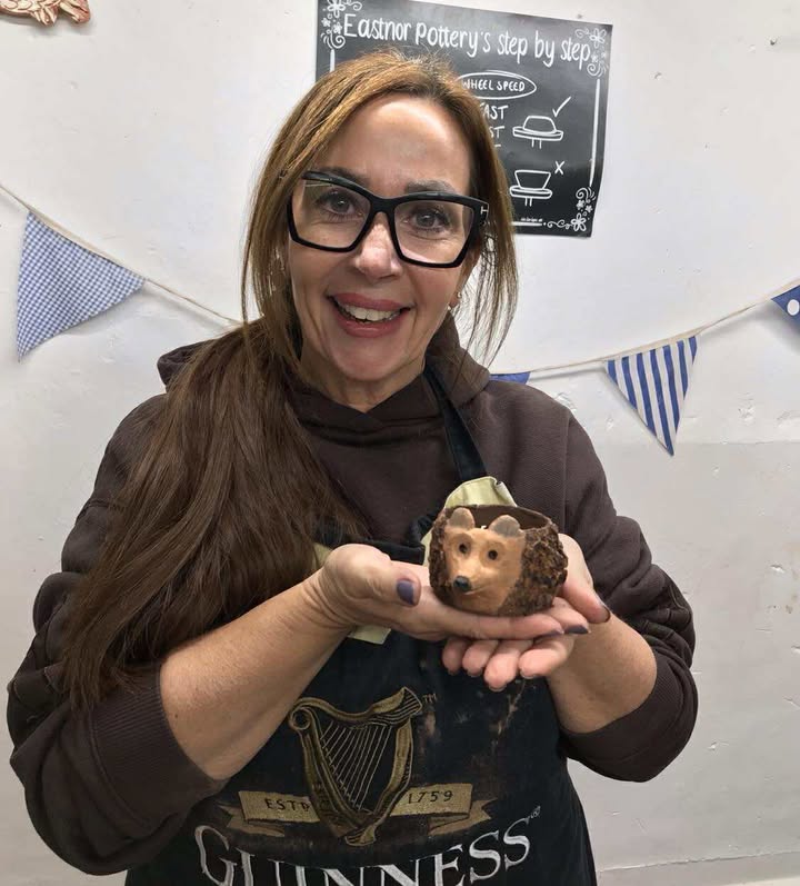A lady with brown hair and a long brown ponytail holding the hedgehog pot she has made. She is holding it in the palm of her hand and smiling towards the camera. The pot has a modelled hedgehog face on the side, painted in light brown and the ears have been painted in grey. The pot has a texture all over it like hedgehog spikes.