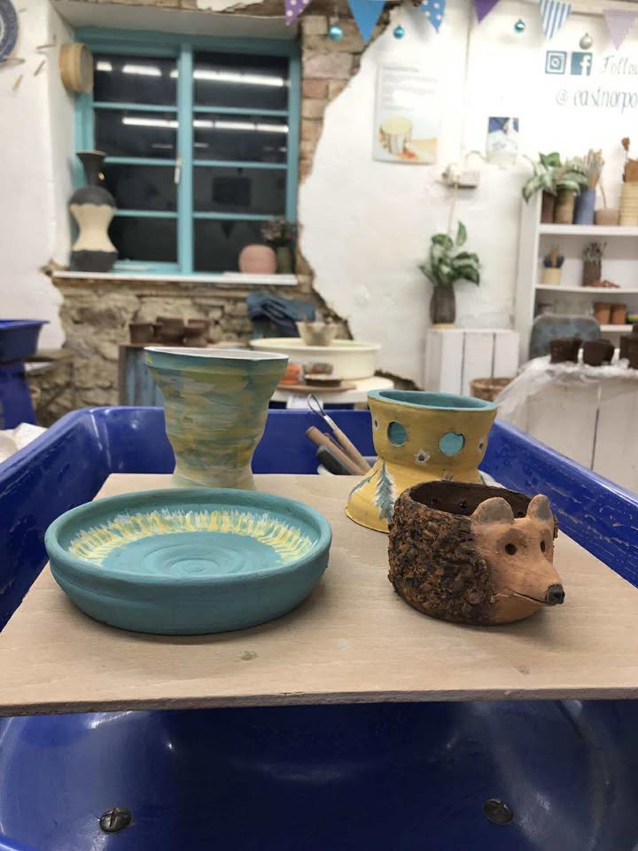 Four finished pots on a wooden square board. A round bowl painted teal with yellow and white feathering around the inside rim, a taller vase with loosely brushed teal, yellow & white, a hedgehog pot and a yellow pot with holes pierced in and feathers painted on the outside.