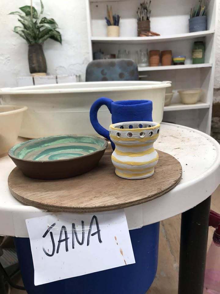 Three finished pots on a round wooden board. A bowl with a green swirl inside, a dark blue mug and a smaller pot with yellow stripes and round holes cut in the rim. There is a piece of paper and it say the name 'Jana'.