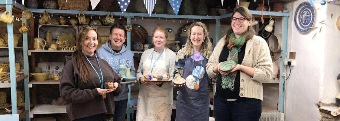 Five happy pottery course participants ! man and 4 women) stood in a row holding pots they made over the weekend. Pots include a pot modelled into a hedgehog,, a bowl with a green swirl and a splattered bowl of yellow and green.