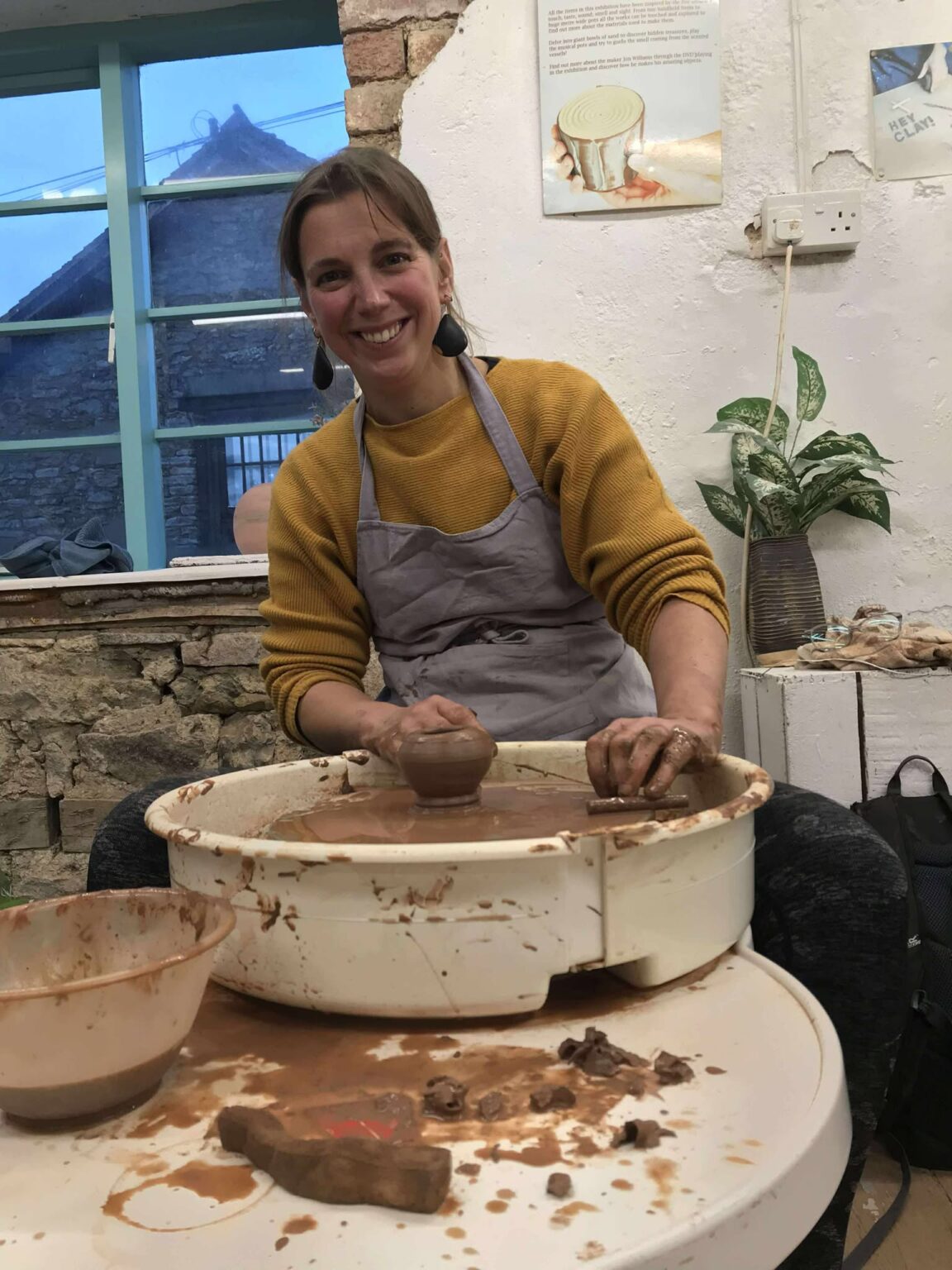 claire from wild cow diary sat on a potter's wheel at eastnor pottery. she is wearing a light grey apron over a mustard coloured cardigan with sleeves rolled up. she is smiling at the camara and has a freshly thrown terracotta clay egg cup on the wheel head in front of her