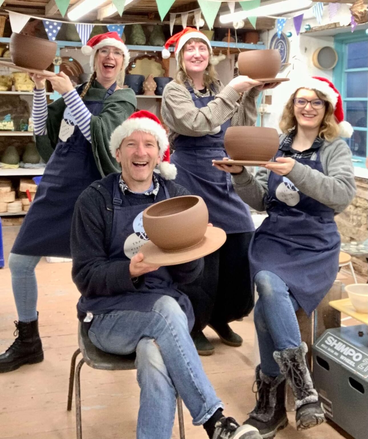x4 staff members at eastnor pottery christmas 2025 all looking at the camera wearing santa hats and holding a brown terracotta pot each on a round board