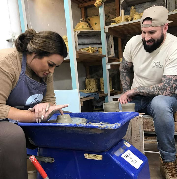 A young couple. A man and a woman. The woman is sat at the pottery wheel, she has very long nails and is concentrating intently. The young man is sat on stool by her side. He is wearing a baseball cap turned backwards and is smiling at her making a brilliant pot.