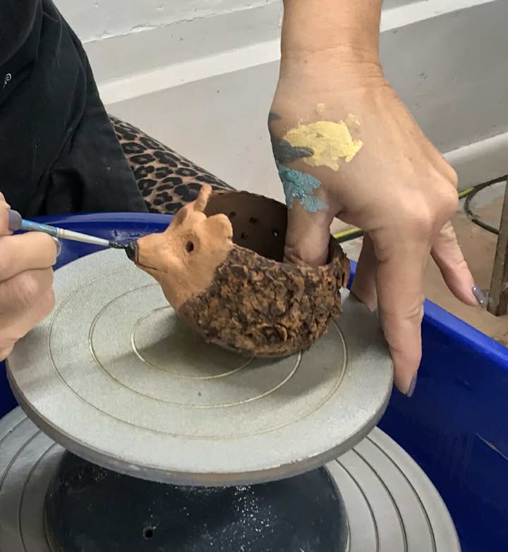 The clay hedgehog from the previous photos is having it's nose painted black with a small art brush. The maker is slightly tipping up the pot. They have paint on their hands.
