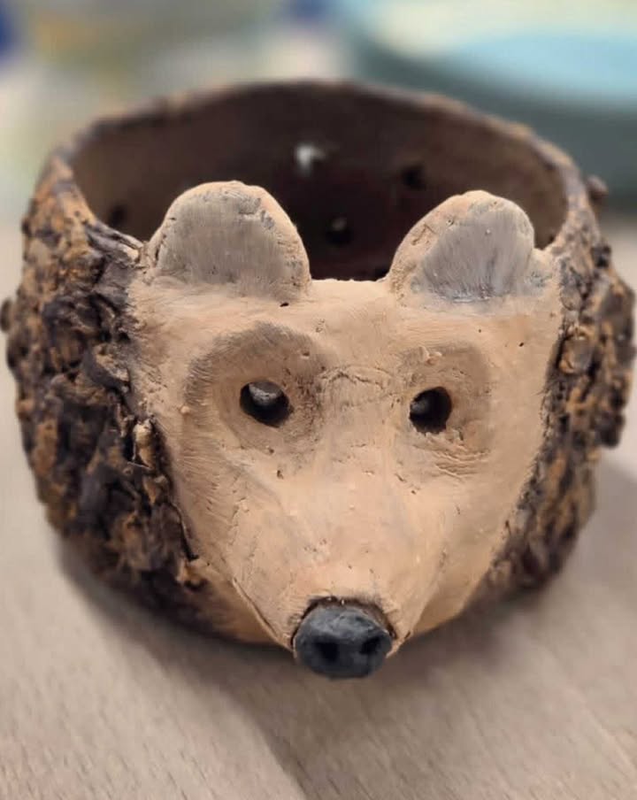 A close up photo of a clay bowl that has been hand modelled into the shape of a hedgehog. It has a light brown modelled face with painted black nose and grey ears. The bowl shape itself has been pierced with holes.