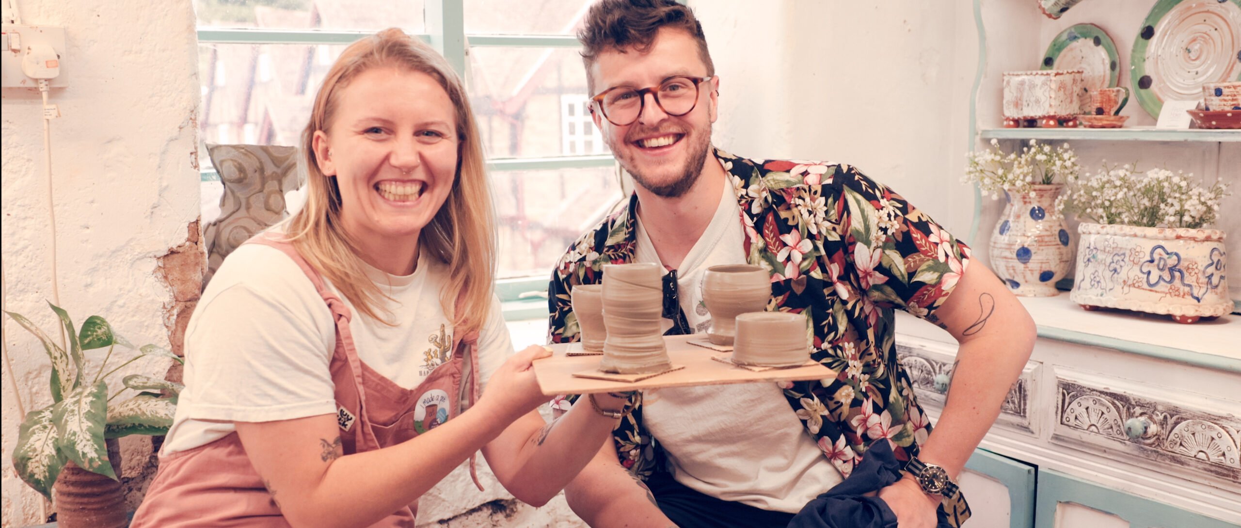 A young couple, a man and woman, sat next to each other, they are both smiling very happy at the camera. The woman is holding up a board of freshly made pot.