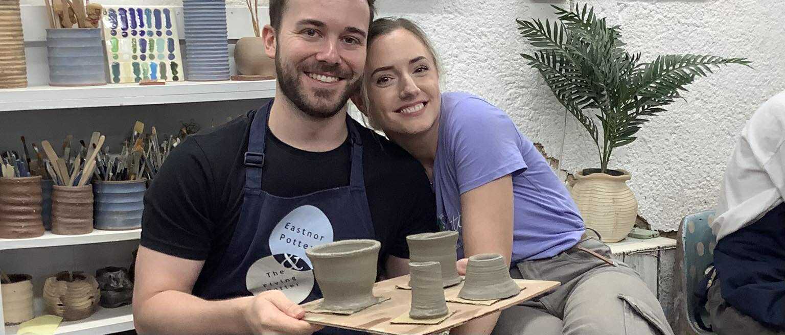 A happy young couple, a man and a woman, sat closely together and leaning their heads in towards each other. They are smiling at the camera. The young man is holding up a wooden board containing 4 freshly made pots.