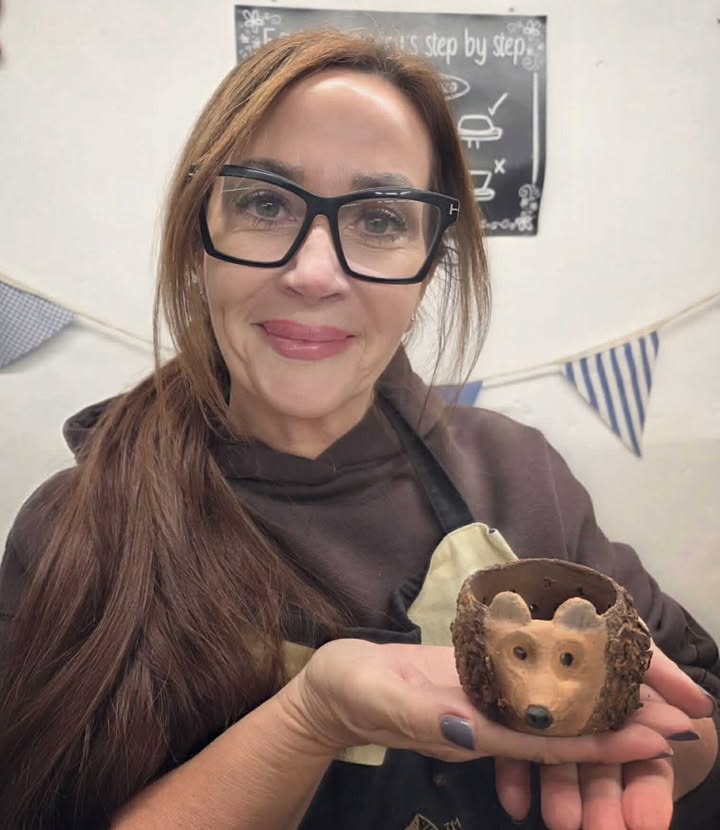 A lady with long dark brown hair and black glasses is holding up a clay hedgehog. It is sitting in the palm of her hand.
