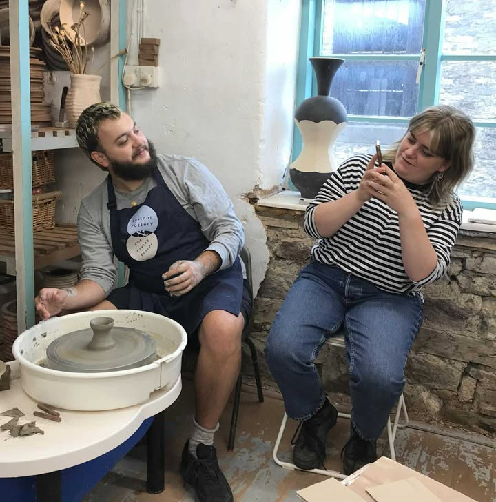Making memories. a young couple in the Eastnor Pottery studio. The man is sat at a potter's wheel and has just made a tall pot, which is on the centre of the wheel. The young lady is sat next to him and is leaning back slightly to take a photo on her phone.