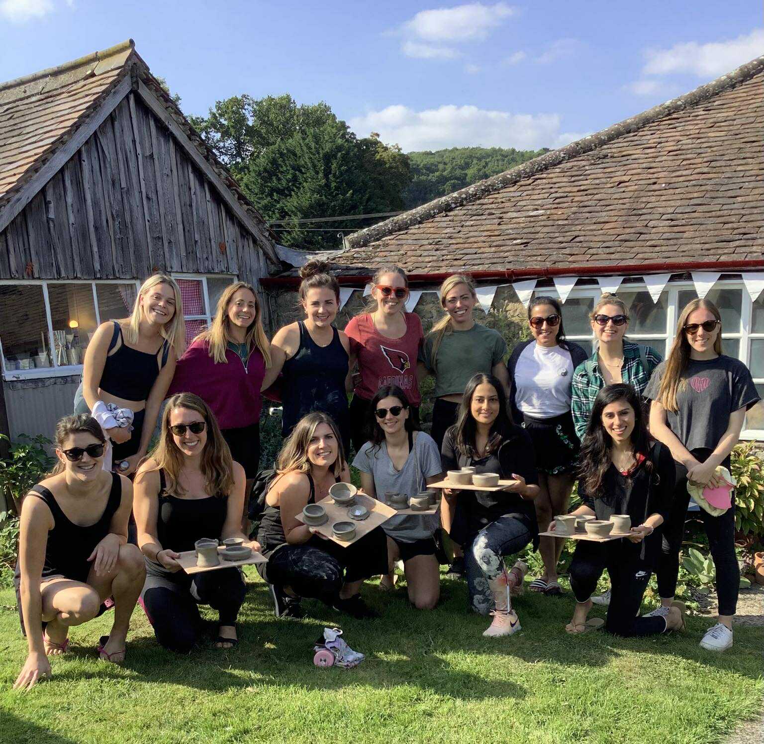 group of x16 women at eastnor pottery in two lines, one line kneeling on the ground, the other line stood behind. It's a sunny day and most of the girls are wearing sunglasses and holding their boards of freshly made pots created on the potter's wheel. It's a pottery hen party.