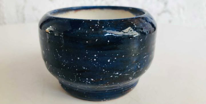 A shiny, fired and glazed finished pot. The glaze is dark blue and black with white splatters and flecks. It is glazed white inside.
