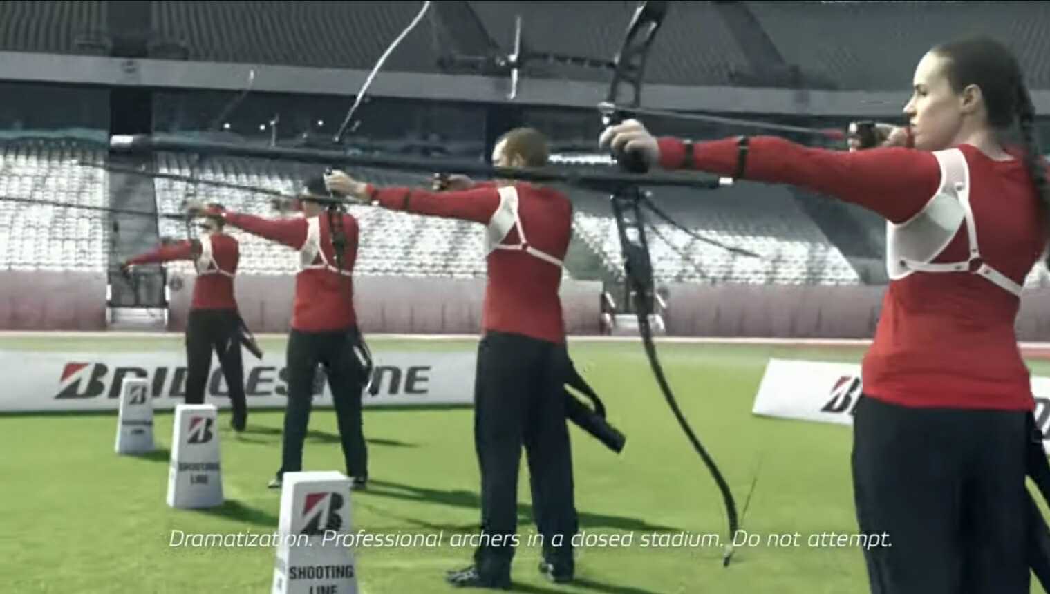 A picture of a row of 4 archers in an empty sports stadium. They are wearing red sports tops and black trousers. The arccers are standing in a row with their arrows poised. Caitlin is at the fornt of the photo.