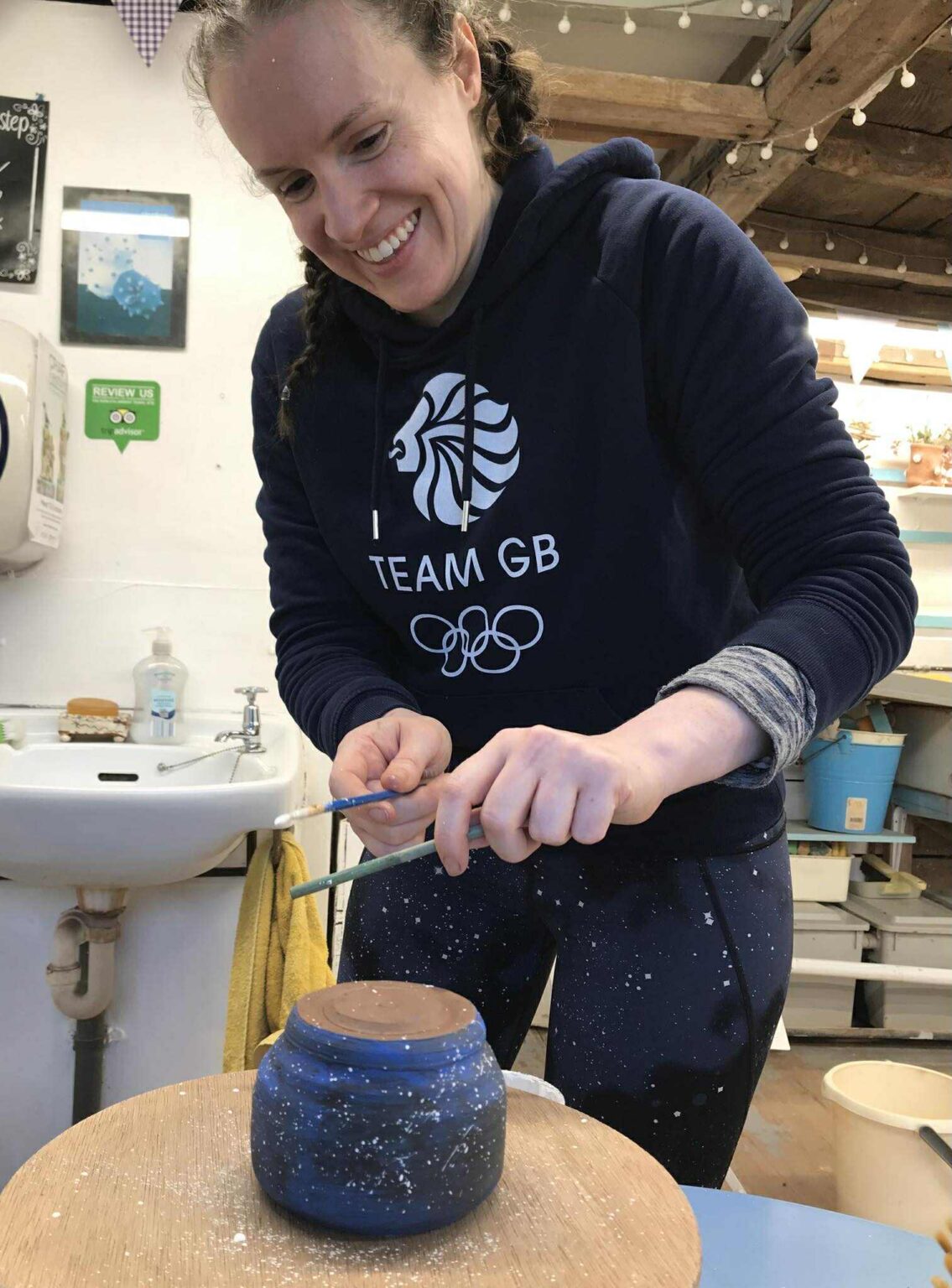 A young women wearing an official olympics sports hoody that says 'Team GB'. She is smiling and looking down at her po that she is decorating.