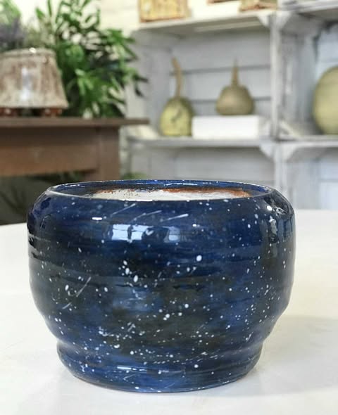 A shiny, fired and glazed finished pot. The glaze is dark blue and black with white splatters and flecks. It is glazed white inside. The pot is sat on a white table at Eastnor Pottery.