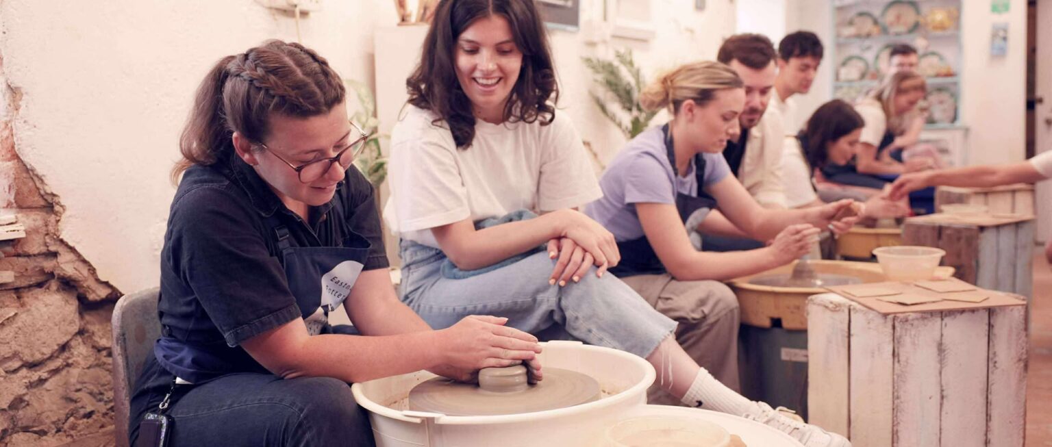 Two young women taking part in the Introduction to the potter's wheel class. One of the women has a disc of clay sat at the potter's wheel. The other is sat to the side, smiling.There are six other people at the potter's wheels alongside in a row.