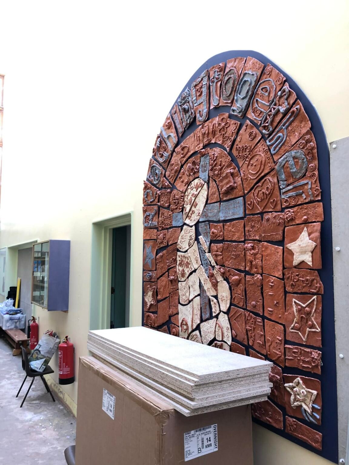 Collaborative mosaic style tile panel made by all the children at ivington primary school herefordshire and jon the potter from eastnor pottery. Each child has created a tile each. The whole panel is terracotta with small areas of blue and white. It is in the shape of a broad arched window. The words Reaching together fill the arch.