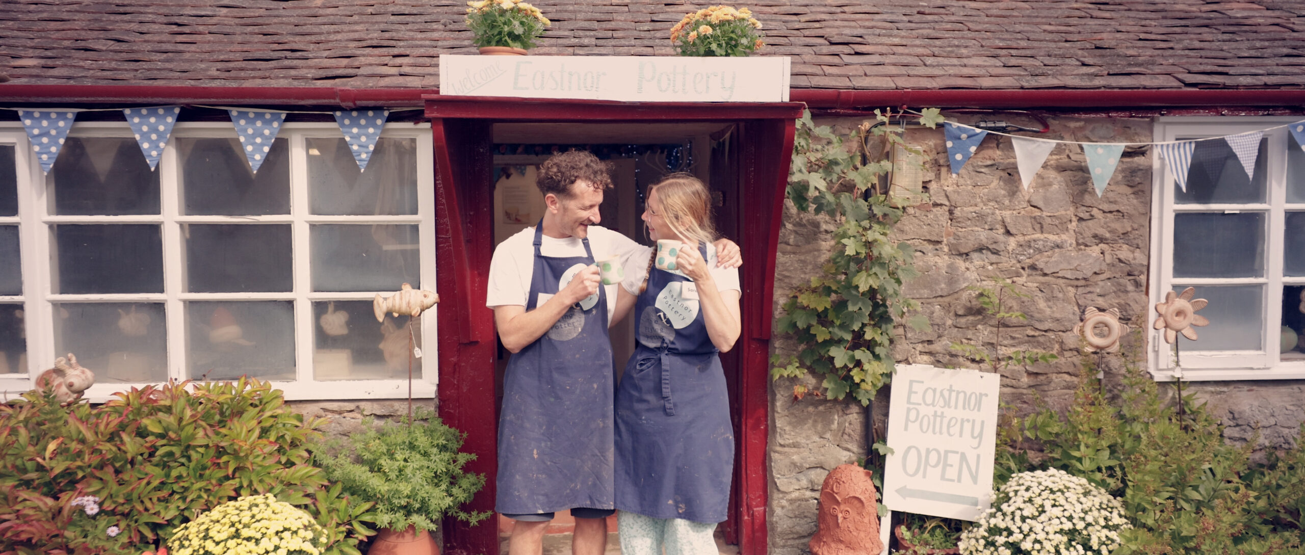 A couple, stood in the doorway with a sign above their heads that say 'Eastnor Pottery', there is an A-board sign next to them too.They are both wearing navy blue aprons and are holding a mug of tea each. They are looking at each other and are smiling.