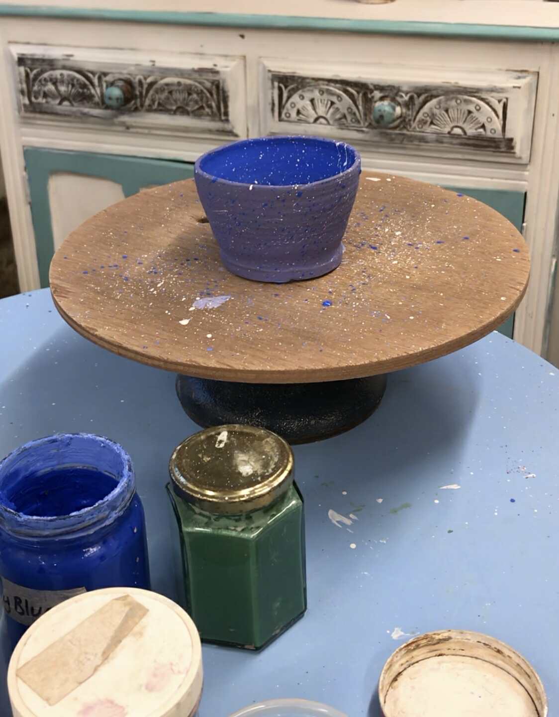 A blue painted pot with white splatters. The pot is on a circular decorating turn table. There are some pots of paint on the table next to it.