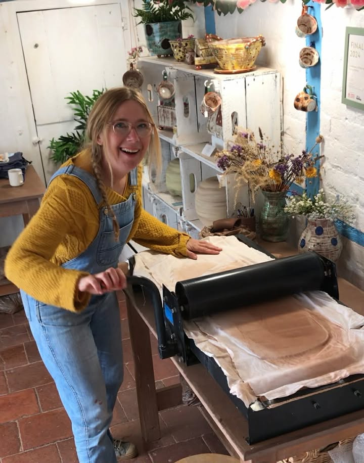 A very happy lady in blue dungarees and a yellow jumper. Turning a large handle of a table top slab roller. The clay is being flattened under a large roller in between a white cotton sheet.