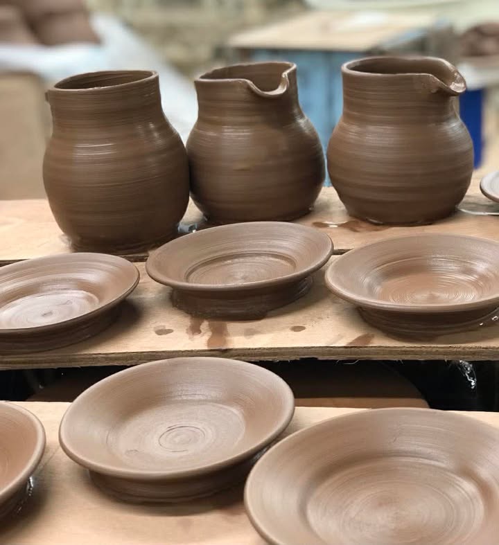 Three freshly thrown terracotta jugs with no handles and six small plates in front.