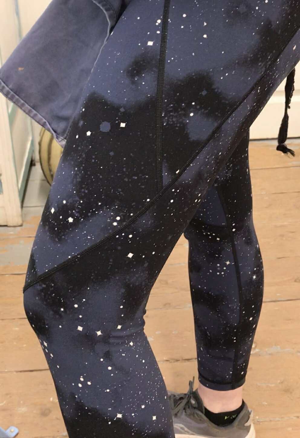 Showing off a pair of leggings Caitlin is wearing. They are dark blue and black. They look like outer space. They are splattered all voer with white stars looking a bit like a galaxy.