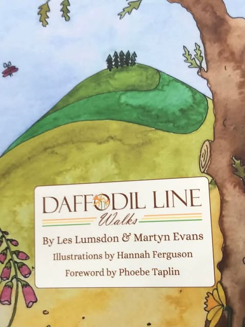 The front cover of a book which says Daffodil Line Walks by Les Lumsdon and Martin Evans. Illustrations by Hannah Ferguson. Forward by Phoebe Taplin. There is a simple and stylised illustration of a hill with three shades of green, in stripes going up the hill and there are 5 trees on the top that look like pine trees. There is a stem of drawn and painted foxgloves to the front left and a simple shaped butterfly above. There is the side of an oak tree and the sky id light blue.