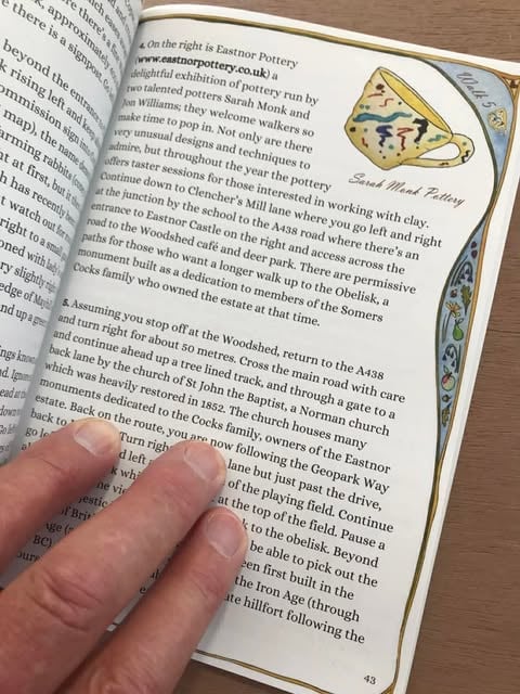 Page 43 of an open book called Daffodil LIne Walks by Les Lumsdon and martyn Evans. Three fingers in the bottom left hand corner are holding the page open. The text suggests walkers should call into eastnor pottery during their hike to view the ceramic work of jon williams and sarah monk. In the top right corner of the image is a hand drawn illustration of a yellow cup with green and blue squiggles on it with the words sarah monk pottery underneath