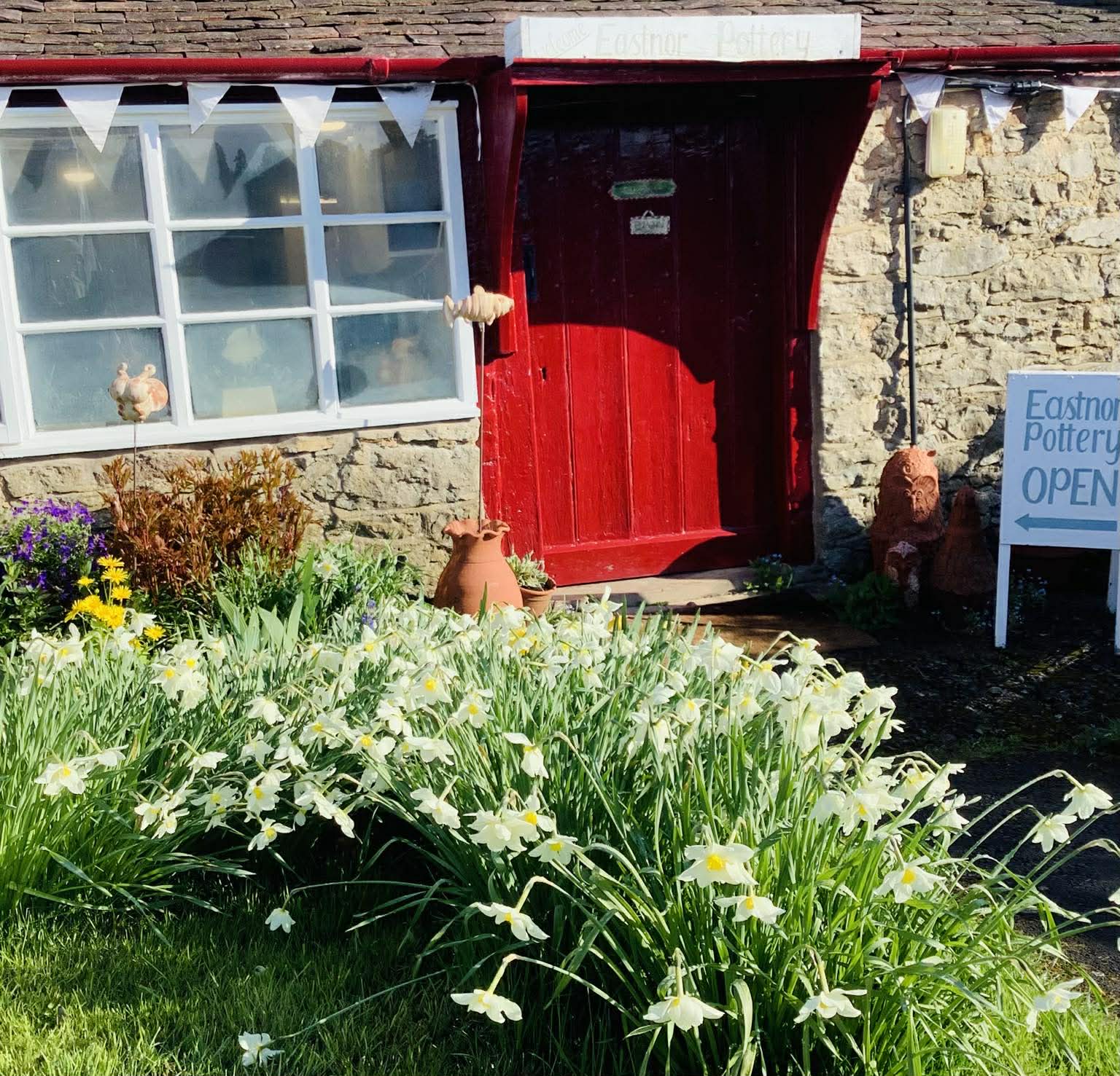 The red painted wooden doorway of Eastnor Pottery on a sunny spring day. The garden is full of daffodils. The Pottery building is made of stone and looks very old, there is white bunting decorating the frontage.