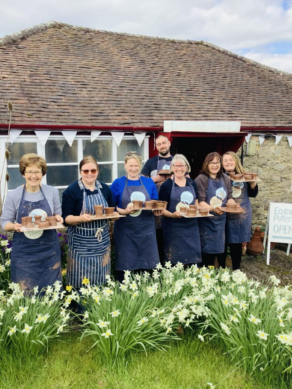 A row of 6 ladies and a taller man stood behind. They are outside in front of Eastnor Pottery, which is an old stone, single story cottage. The people are wearing aprons and holding boards of pottery they have made. Their is an abundance of daffodils in front of them, growing out of the lawn.
