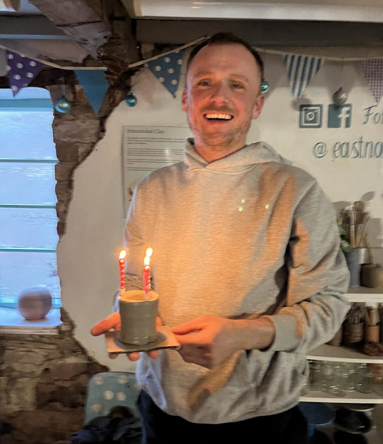 birthday pottery participant steve laughing at the camera and holding a wet pot he's made on the potter's wheel. The pot has x2 lighted birthday cake candles. The light from the flames lights up the front of his beige hooded top