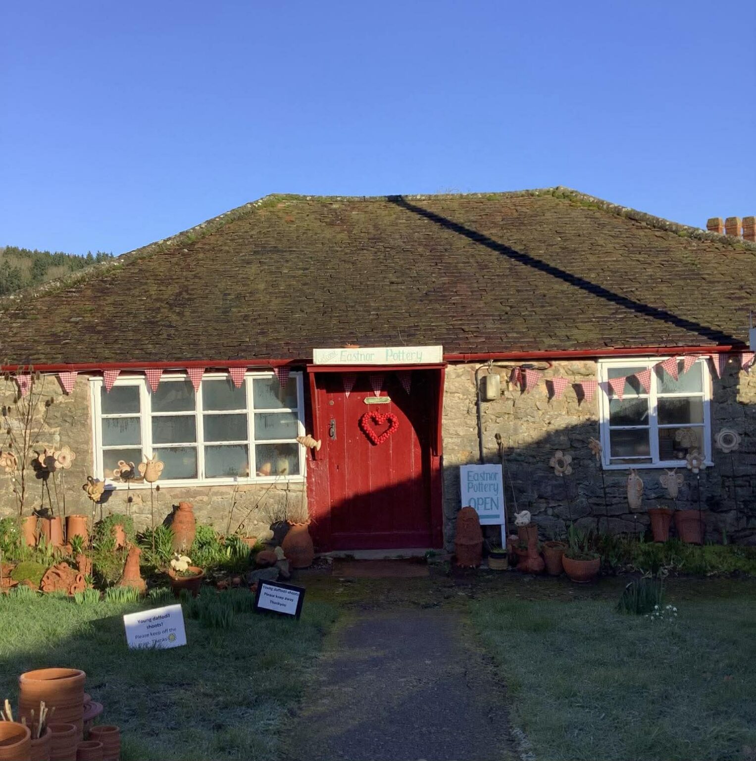 The front of Eastnor Pottery. A single storey country cottage with a bright burgandy door. A single rectangular window with twelve glass panes sits to the left. A deep diagonal shadow falls across half the image covering most of the garden.