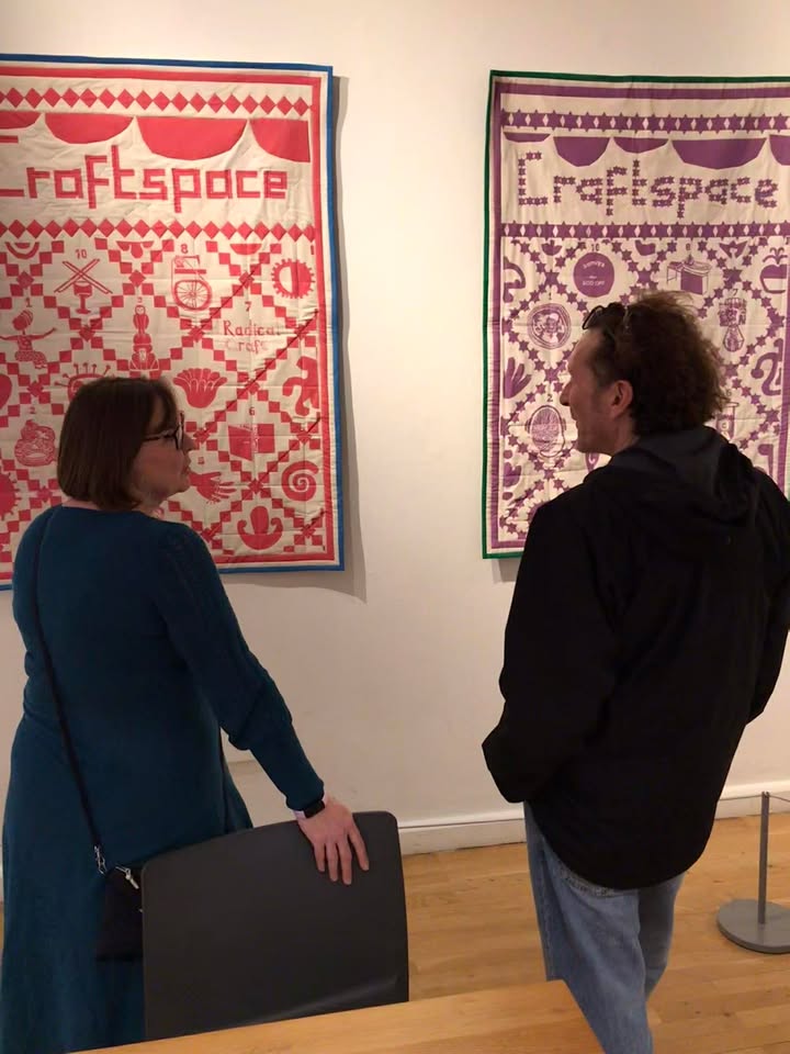Background: Two quilts hanging on the wall at Midland Arts Centre, red one on the left and purple one on the right. The quilts were commissioned to celebrate 40 years of craftspace. Two figures in the foreground are silhouetted, facing each other and in deep conversation. The male figure on the right is ceramic artist Jon Williams and the female figure on the left is a senior member of the craftspace team.