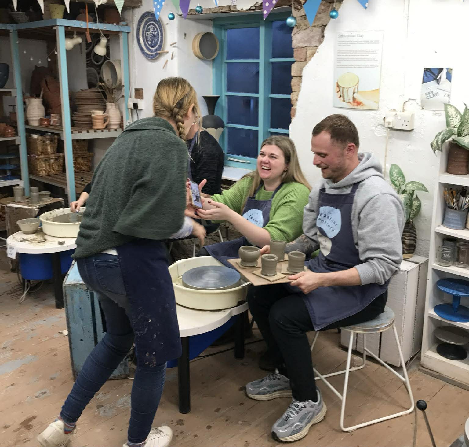laughing couple sat at the potter's wheel at eastnor pottery. The man on the right is holding a board of four freshly thrown grey pots. Sarah Monk from Eastnor pottery is showing the couple the photograph she has just taken of them