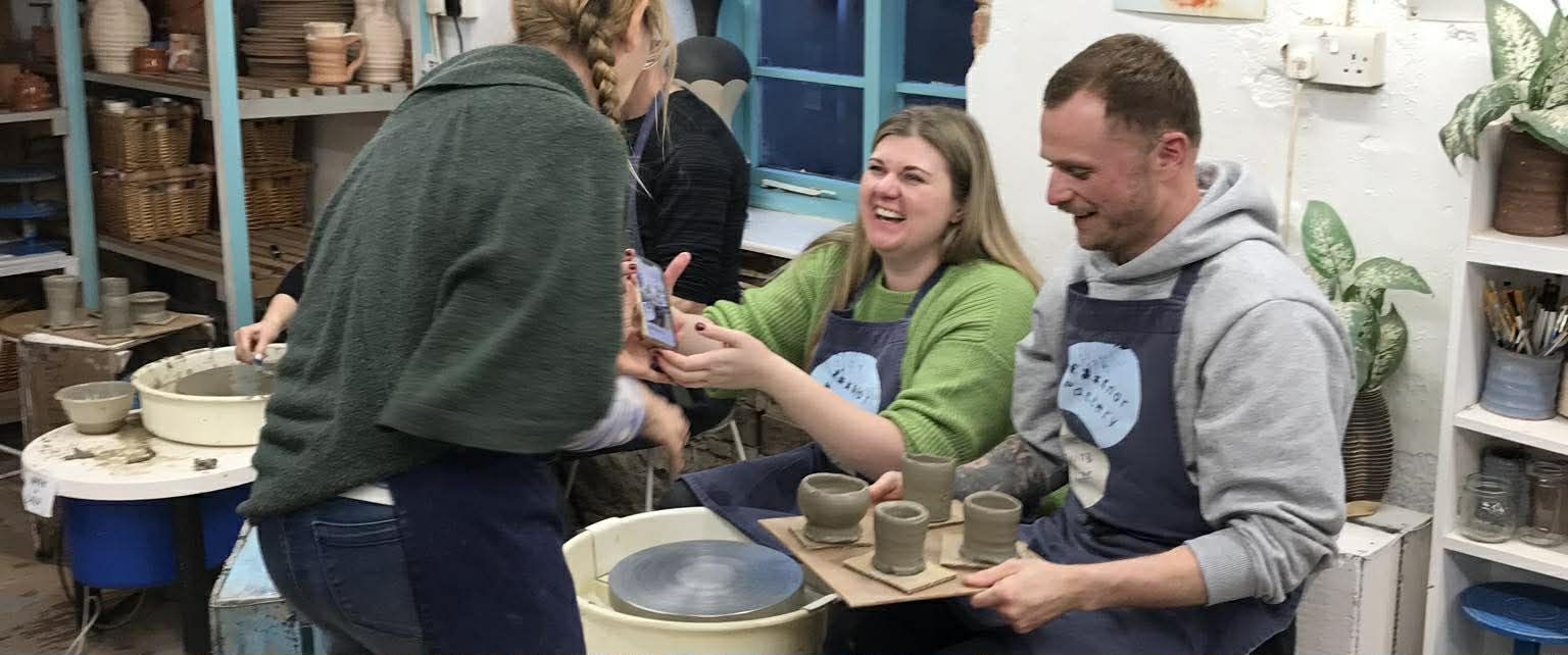 laughing couple sat at the potter's wheel at eastnor pottery. The man on the right is holding a board of four freshly thrown grey pots. Sarah Monk from Eastnor pottery is showing the couple the photograph she has just taken of them