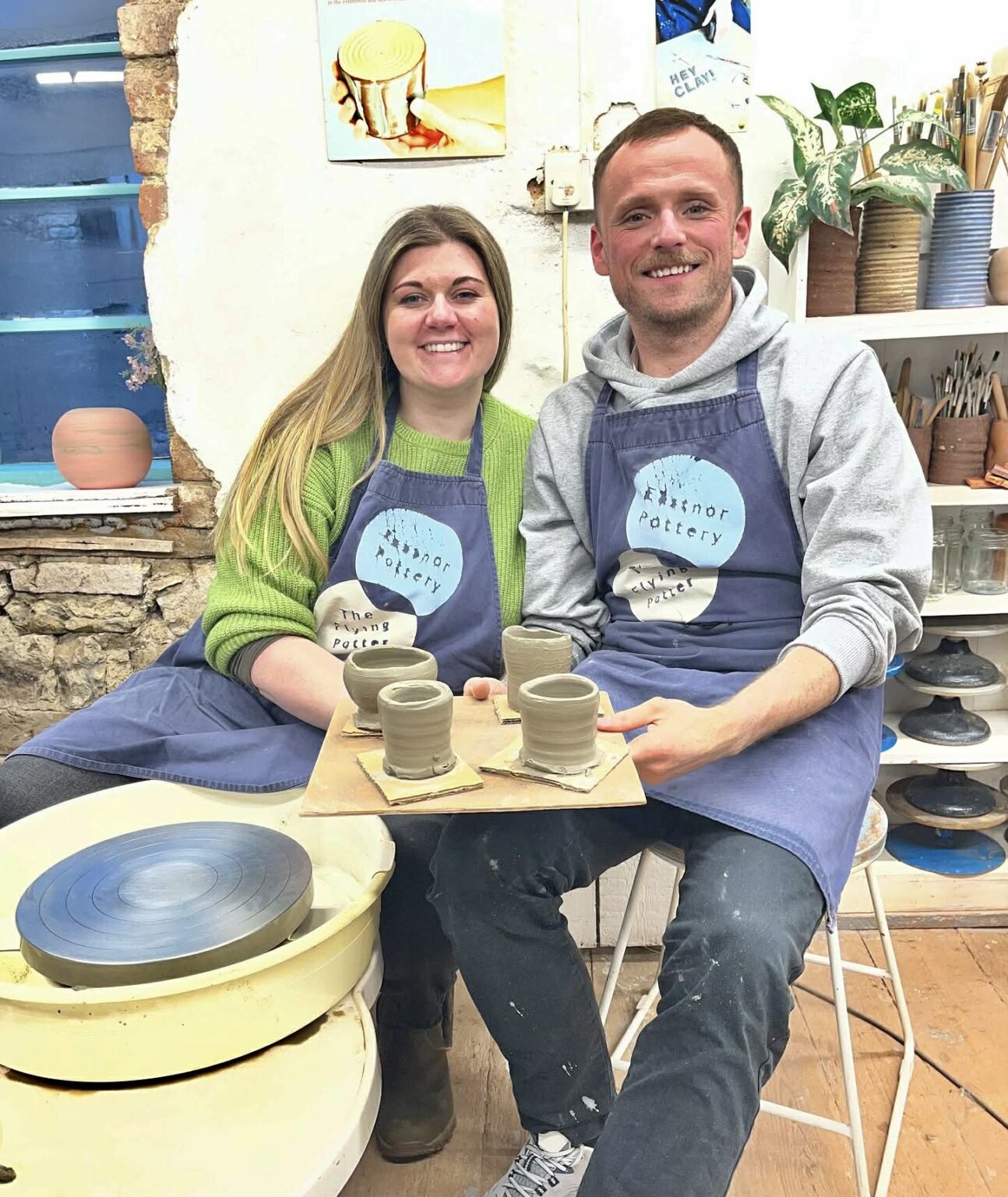 smiling couple looking straight at camera jointly holding a square board full of soft thrown pots made on the potter's wheel at eastnor pottery. The pair are wearing blue aprons with eastnor pottery logo on them. A clean potter's wheel head fills the bottom left hand corner