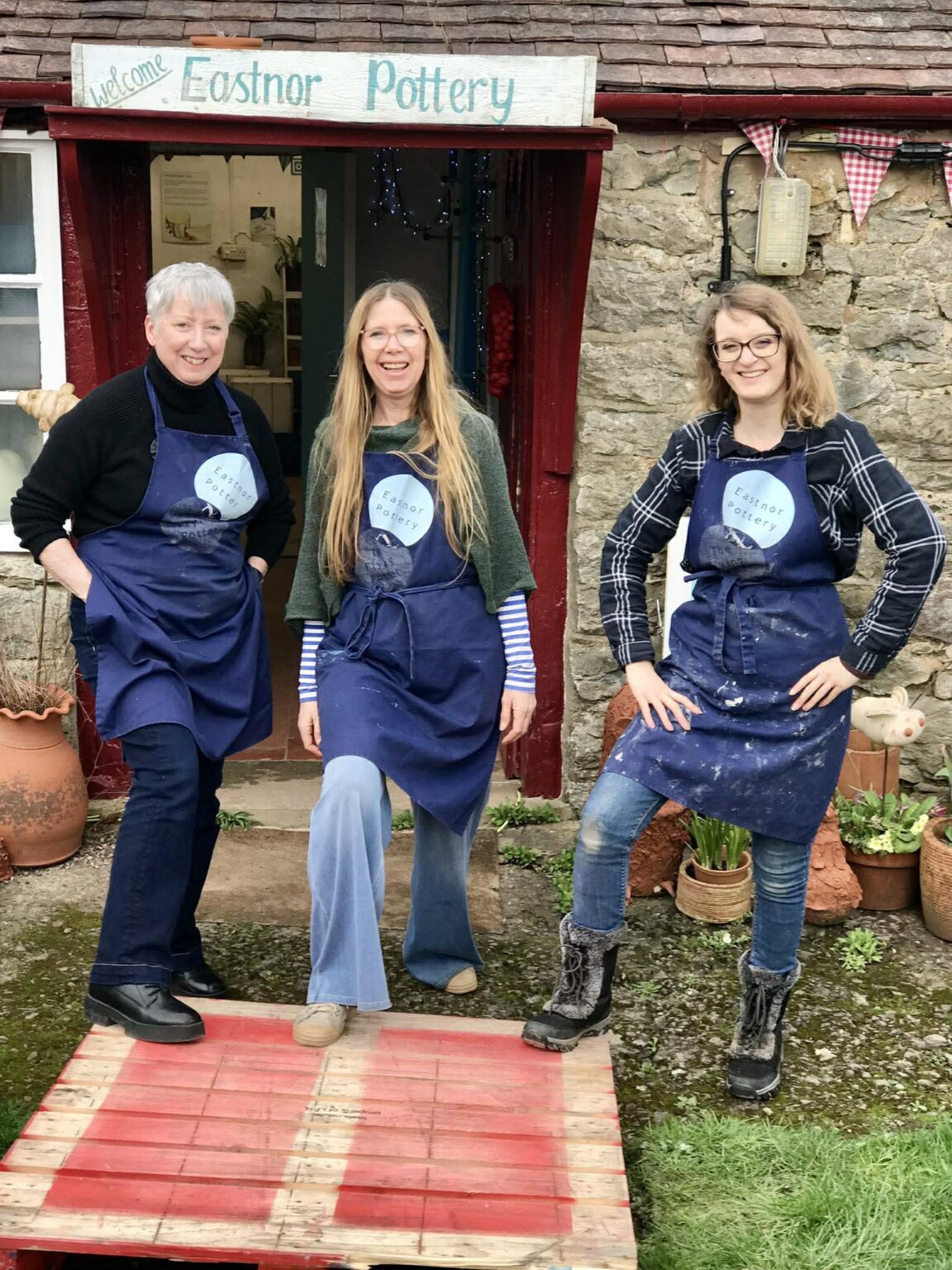 From left to right Sylvia, Sarah and Aimee from eastnor pottery. full length figures in a row. Each woman has a foot on a wooden pallet and is smiling at the camera
