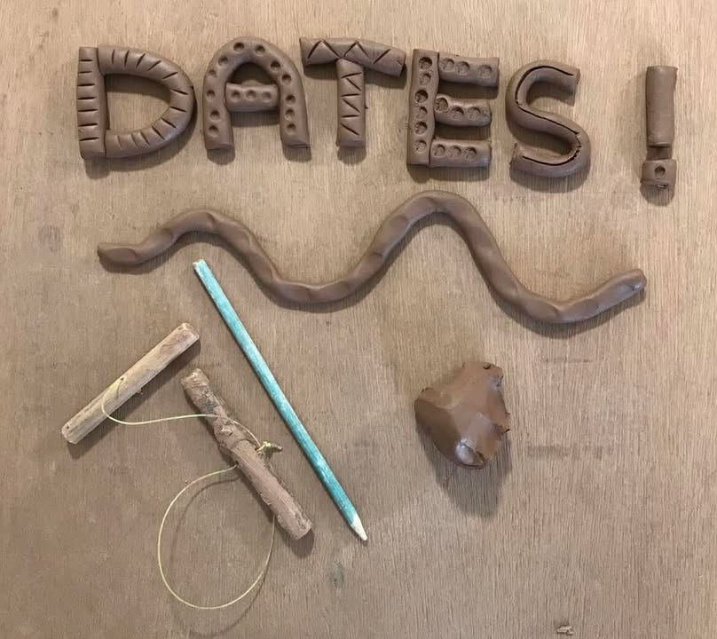 The word DATES with an exclamation mark made out of rolled terracotta clay and indented with dots and stipes. There is a wiggley clay coil underneath the words plus a pottery tool called a cheese wire and a wooden clay modelling stick. It is arranged on a wooden table top