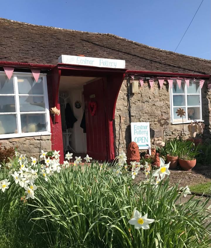 The red front door of Eastnor Pottery within the pottery garden. The door is open on a sunny day with blue sky and there are large bunches of daffodils growing in the grass. 