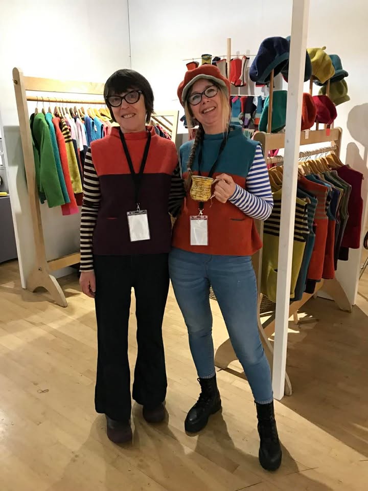 Two ladies standing together with lanyards and wearing similar tank tops but in different colours. They have their arms around each other and they are smiling at the camera