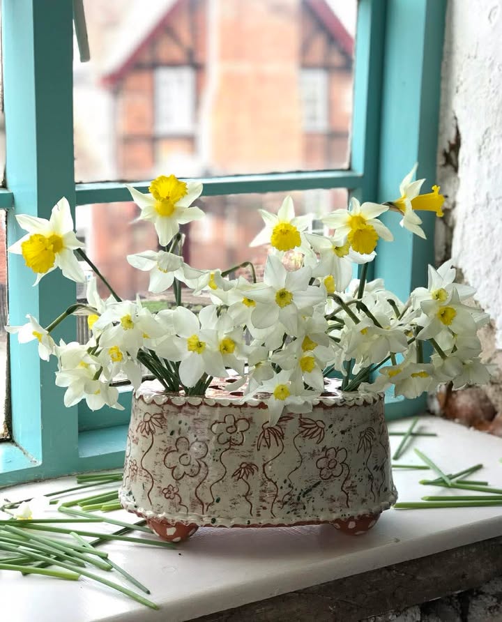 An oval flower brick in cream with etched flowers. It's on a windowsill and is full of fresh flowers. The unwanted cut off stems are scattered on the windowsill.