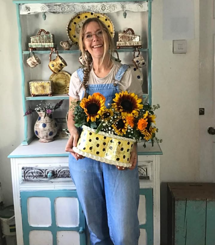 A smiley lady (Sarah Monk) holding a large flower brick. It is yellow with brown  polka dots. It has lots of fresh sunflowers in it. The lady is wearing denim dungarees and has blond hair.
