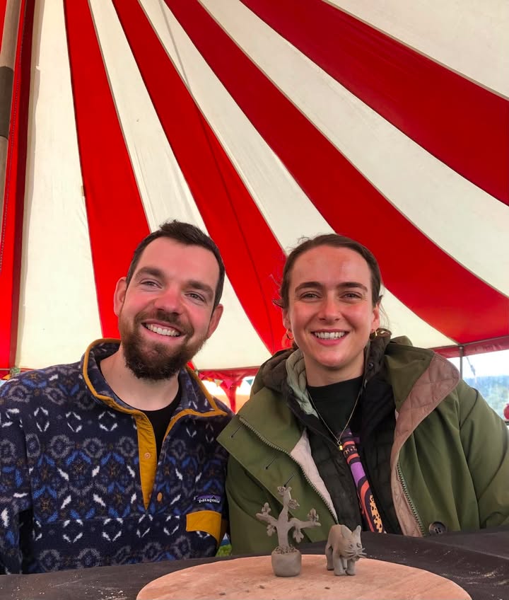 A young couple, man and lady sitting next to each other and smiling at the camera. In front of them on around wooden board are two clay sculptures of a tree and a cat. The couple are sat inside a circular circus tent of red and white stripes.