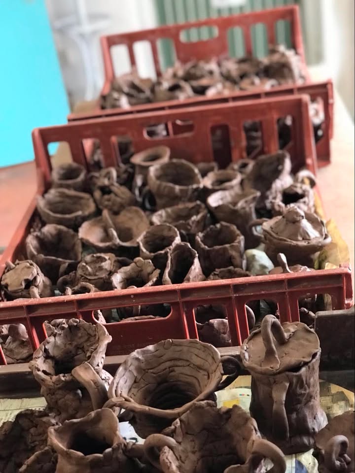 Three plastic bakers crates full of terracotta pots made by school children. They are on a table in a line. Some pots have lids.
