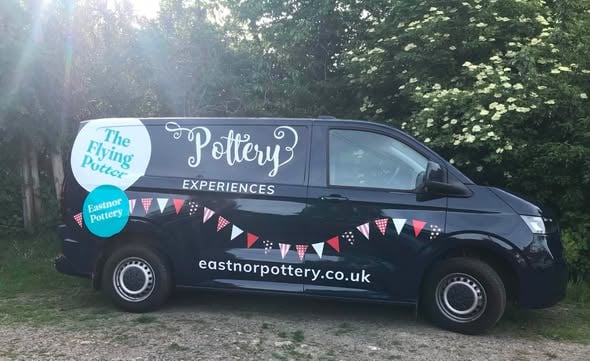 The side of a dark blue VW Transporter van decorated with large transfer stickers there is a swaithe of bunting in red and white, a large white circular sticker that says 'The flying potter' and below it a turquoise circular sticker that says 'Eastnor Pottery' The is large lettering on the side in fancy curly writing that says 'Pottery Experiences'. The website address runs along the bottom that reads 'eastnorpottery.co.uk