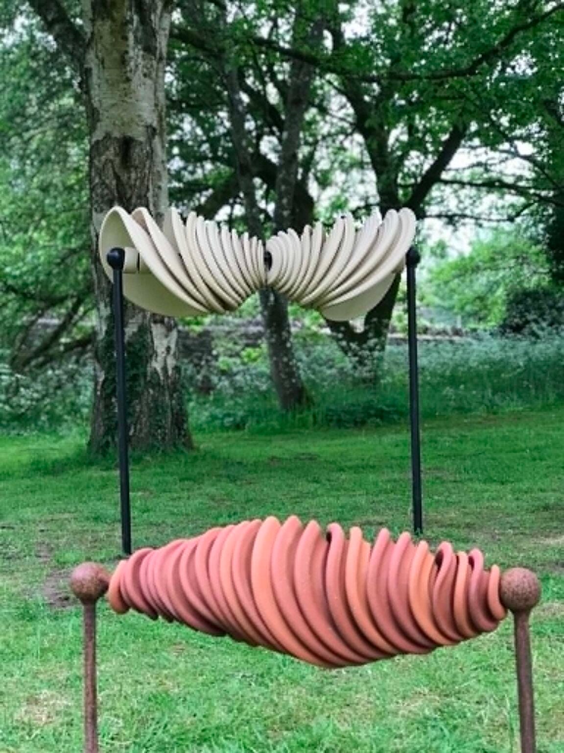interactive percussive ceramic disks made by ceramic artist jon williams. In the forground there is a terracotta arrangement of disks in the shape of a fishes body. In the background a cream coloured horizontal arrangement in the shape of an hour glass on it's side. there are trees in the back ground and the sculptures are situated on a beautiful lawn at fresh air sculpture exhibition in the old rectory in quenington