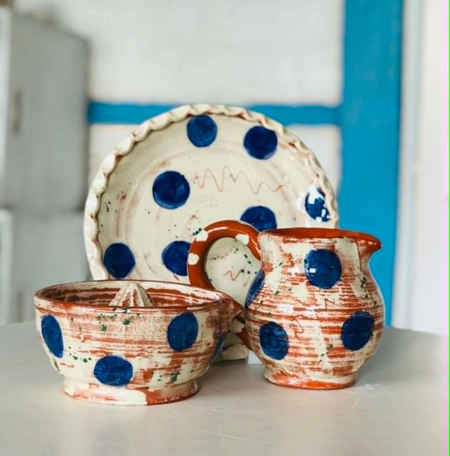 Three pieces of pottery a plate, a juicer and a jug. They are white with large  blue dots with a little bit of terracotta showing through.