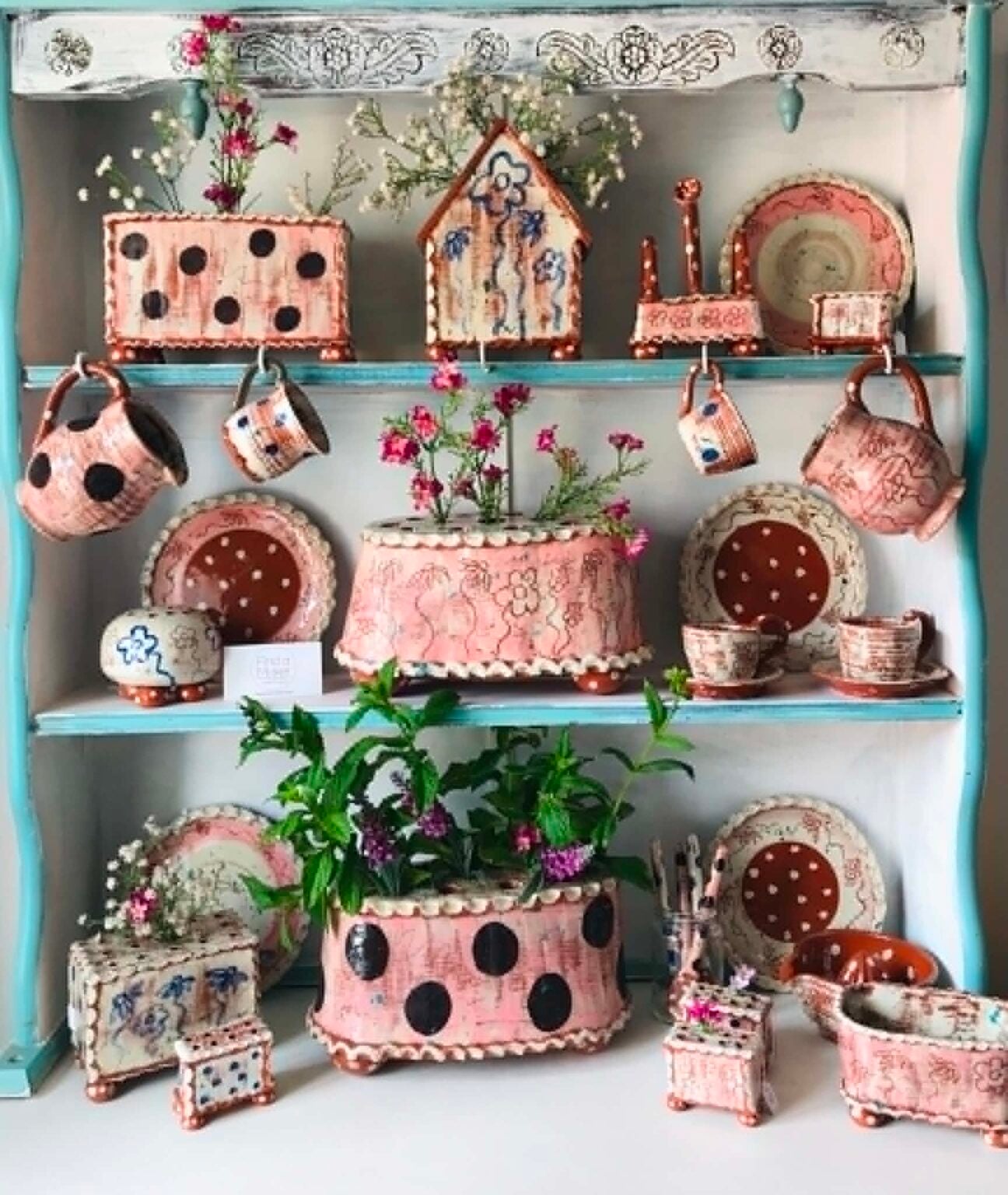 Three shelves of pink and white pottery made by Sarah Monk. There are cups, jugs, plates and flower bricks.