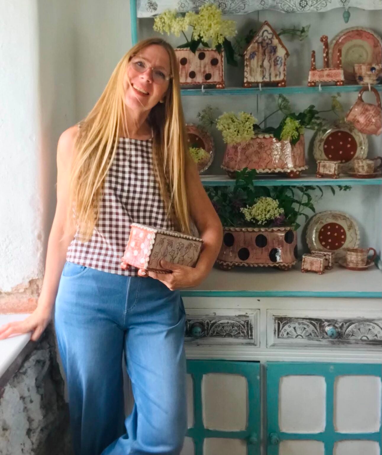 Sarah Monk studio potter stood by a blue and white painted dresser... she has long blond hair , she is wearing pink glasses, blue jeans and a brown and white checked cotton top. She is holding one of her rectangular flower bricks. The dresser is full of pink and white pottery