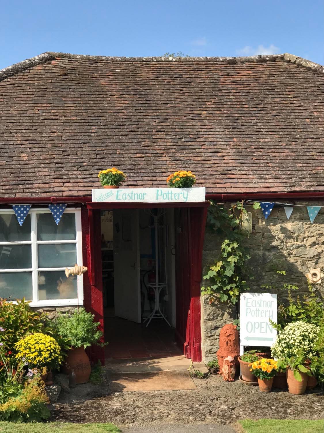 Front on view of eastnor pottery a single storey cottage with large tiled roof. There is a white open sign to the fore and various plants and flowers in pots arranged around the open front door. There is a line of triangular bunting tied to the horizontal gutter.
